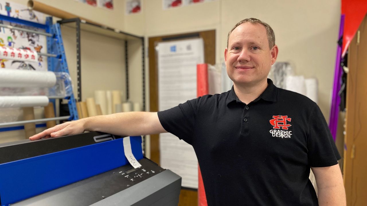 Cedar Hill High School graphic design teacher Aaron Kennedy poses for a picture with the school's wide-format printer. (Spectrum News 1/Lupe Zapata)
